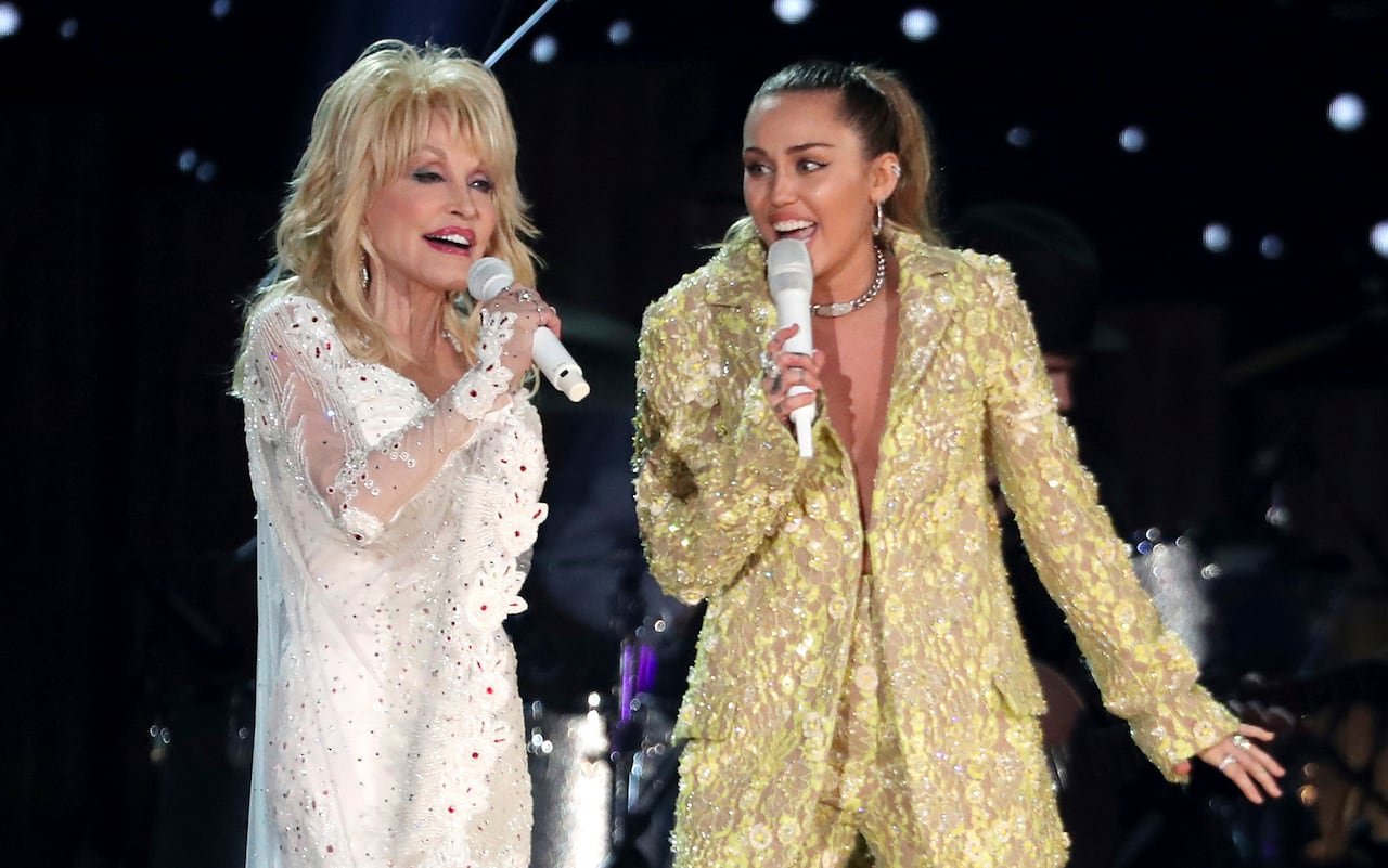 An older lady in a shiny white outfit performs on stage with a young woman in a shiny gold outfit.