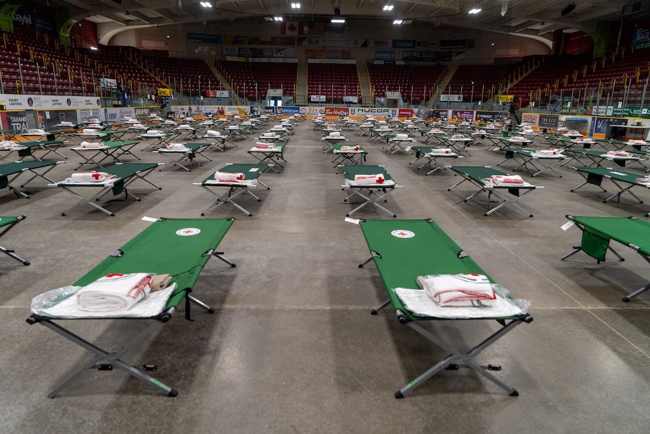 Dozens of green cots are set up on the concrete floor of an arena.