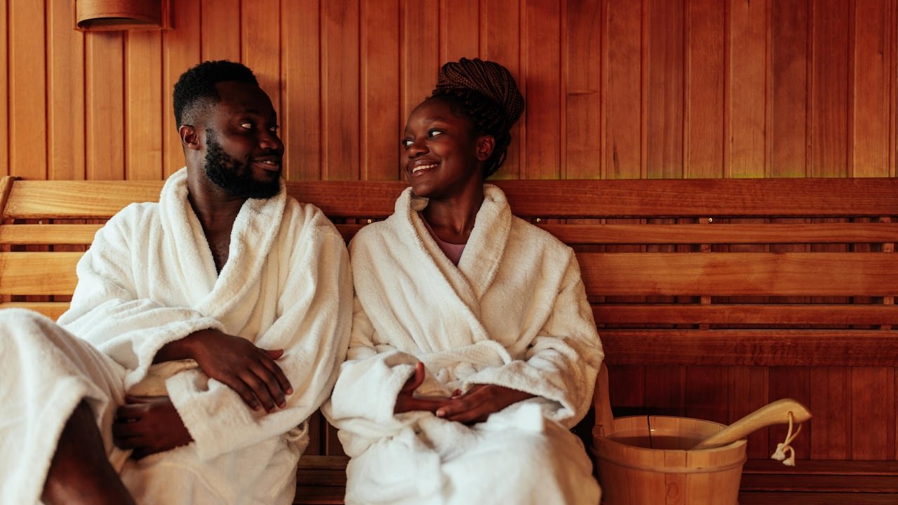 A couple wearing bath robes, sitting on a bench in a wood-panelled sauna, smiling at one another.